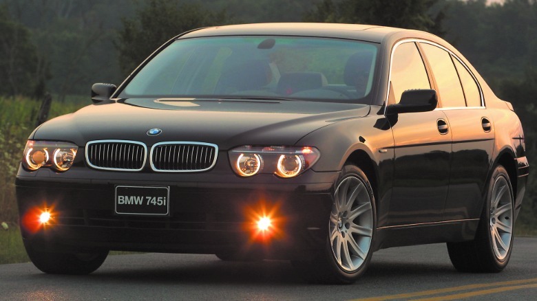 Front three-quarters shot of a black 745i parked on a two-lane road in front of grass and trees at dusk with its fog lights on
