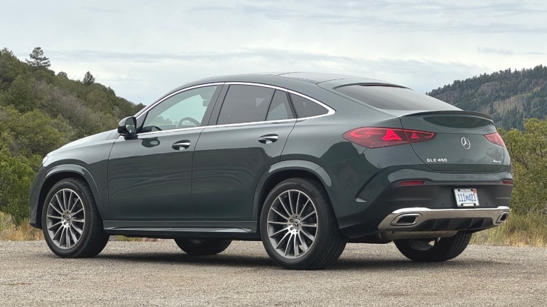 A rear three-quarters shot of a gray GLE450 Coupe parked on gravel with tree-covered mountains and a cloudy sky in the background