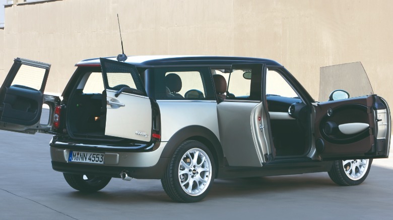 A rear three-quarters shot of a silver Mini R55 Clumban with all of its six doors open parked in front of a tan wall