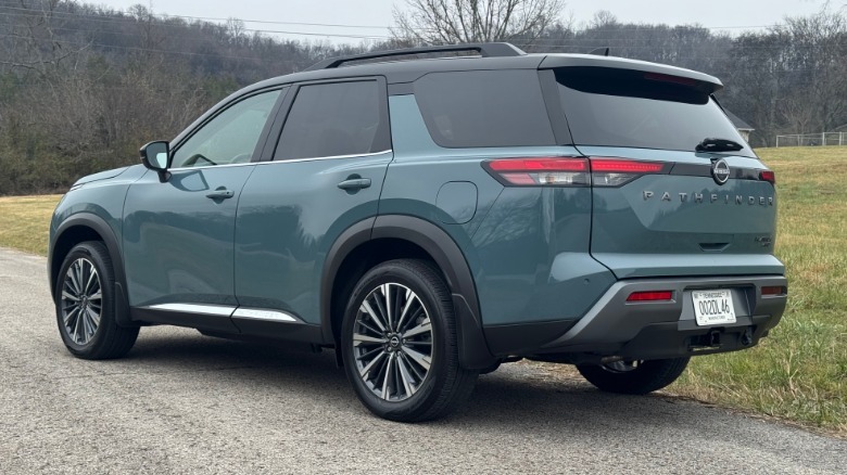 Rear three quarters shot of the teal Pathfinder Platinum parked on asphalt in front of grass and mountains with leafless trees