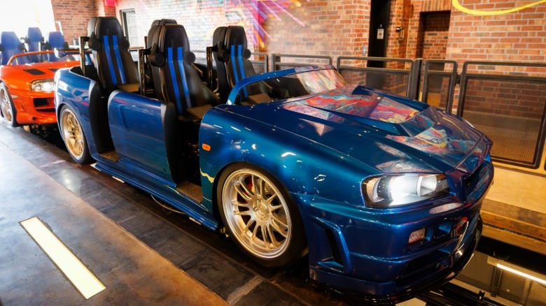 A front three-quarters shot of the blue R34 Skyline GT-R in front of the orange Supra on the ride in front of a brick wall