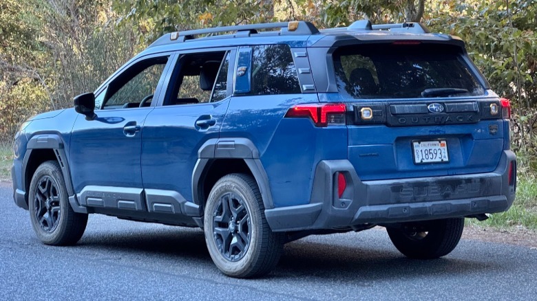 A rear three-quarters shot of the dark blue Outback Wilderness parked on asphalt in front of trees
