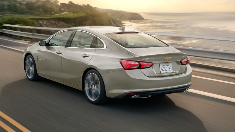 rear three quarters shot of a gold Malibu driving across a bridge over the ocean at sunset in front of green hills