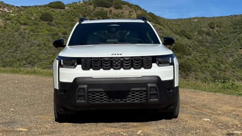 A head-on shot of a white Cherokee parked on gravel in front of a shrubby mountain
