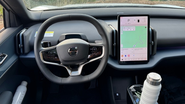 A shot of the driver's POV of the squircle steering wheel and tall center mounted tablet with two white water bottles in cupholders