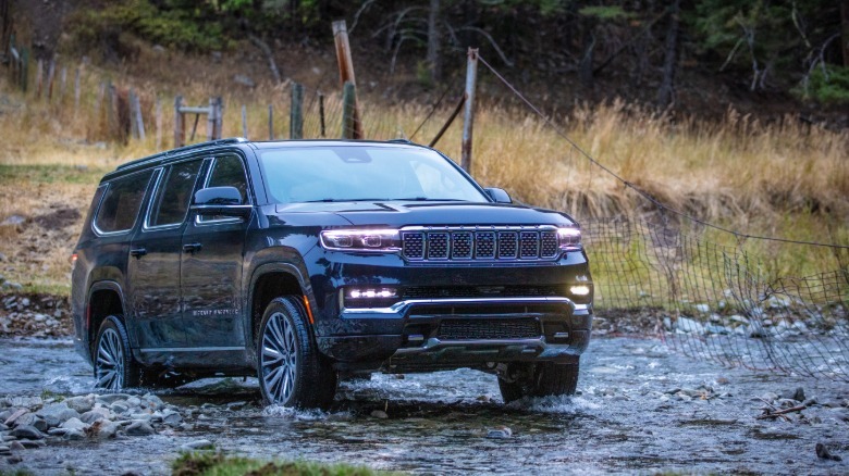 a front shot of a black Grand Wagoneer L driving through a pebbly stream in front of tall grass