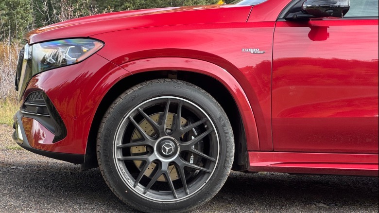 A close-up of the front quarter panel and black wheel of the red GLE53 coupe parked on gravel with some trees and brush visible in the background