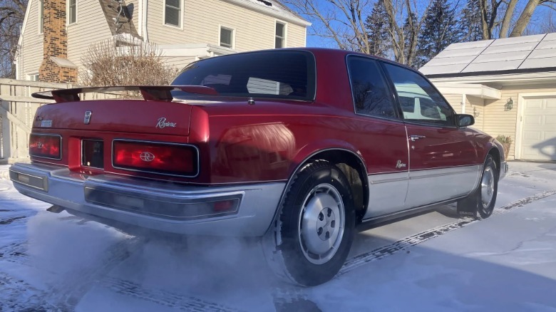 rear three-quarters shot of the burgundy and silver Riviera parked on a snowy driveway in front of a fence and homes with warm exhaust creating steam coming from the exhaust pipes