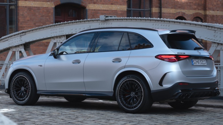 A rear three-quarters shot of the matte silver AMG GLC53 SUV parked on a cobblestone bridge in front of a brick building