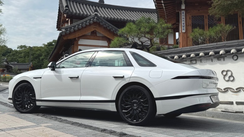 A rear three-quarters shot of a white Grandeur parked on brick in front of an ancient traditional Korean home with black roof tiles